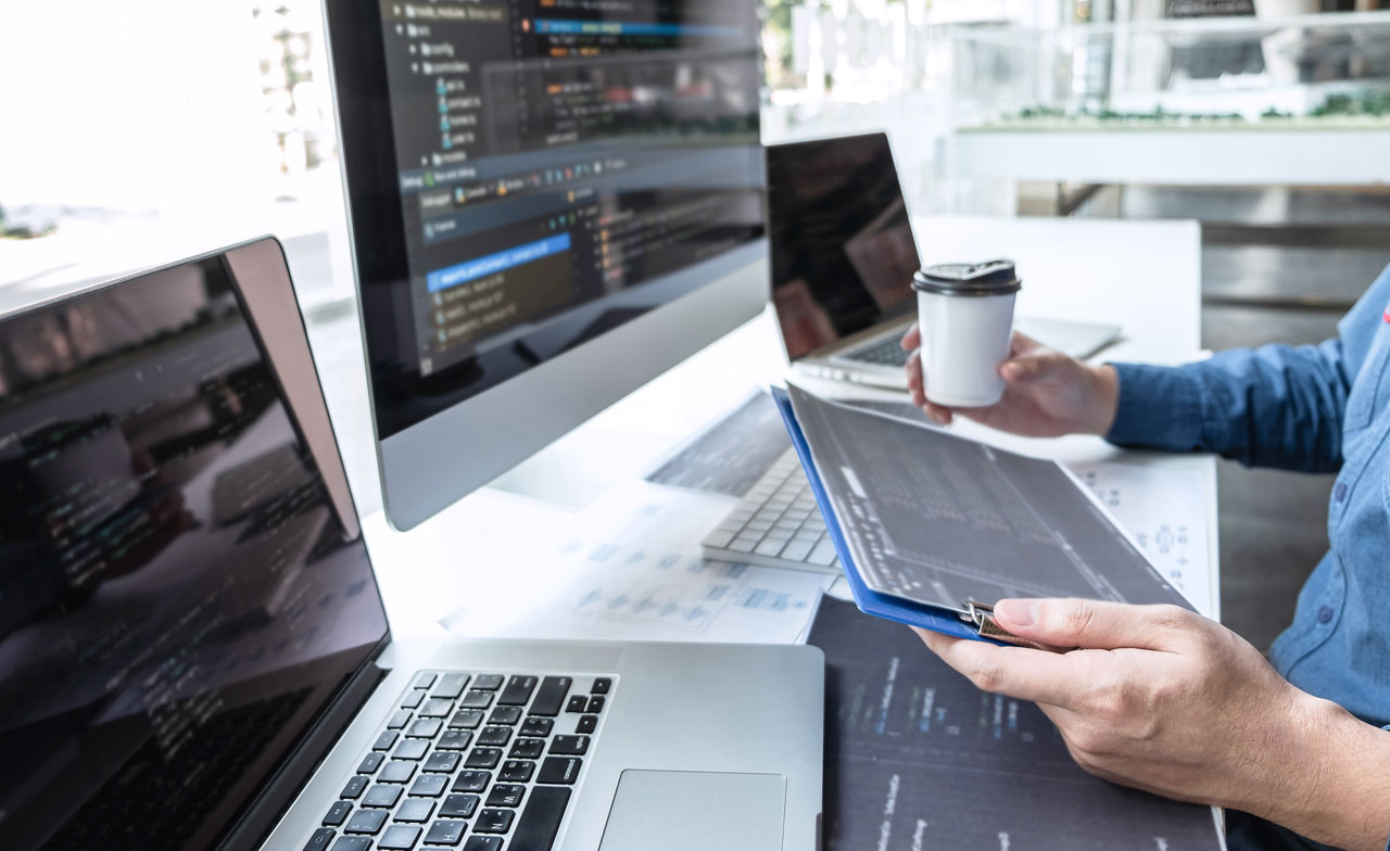Close up of person sat at a desk looking at a clipboard with a laptop and computer screen in the background